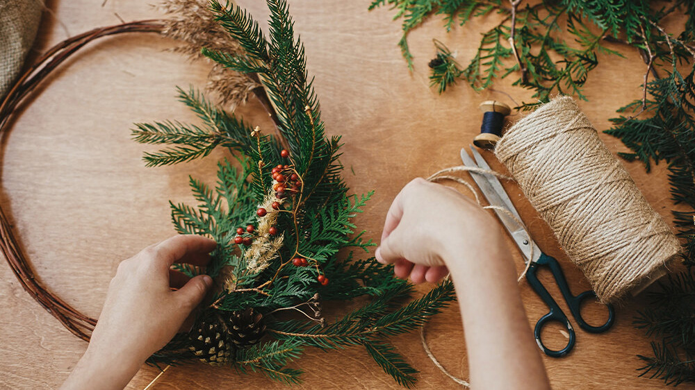 Overhead view of a person making a holiday wreath with twine, pine leaves and holly on a wood table.