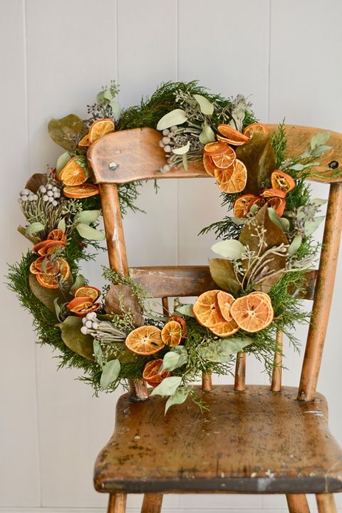 Completed holiday green and orange wreath hanging on the back of a rustic wooden chair with a white wall behind the chair.