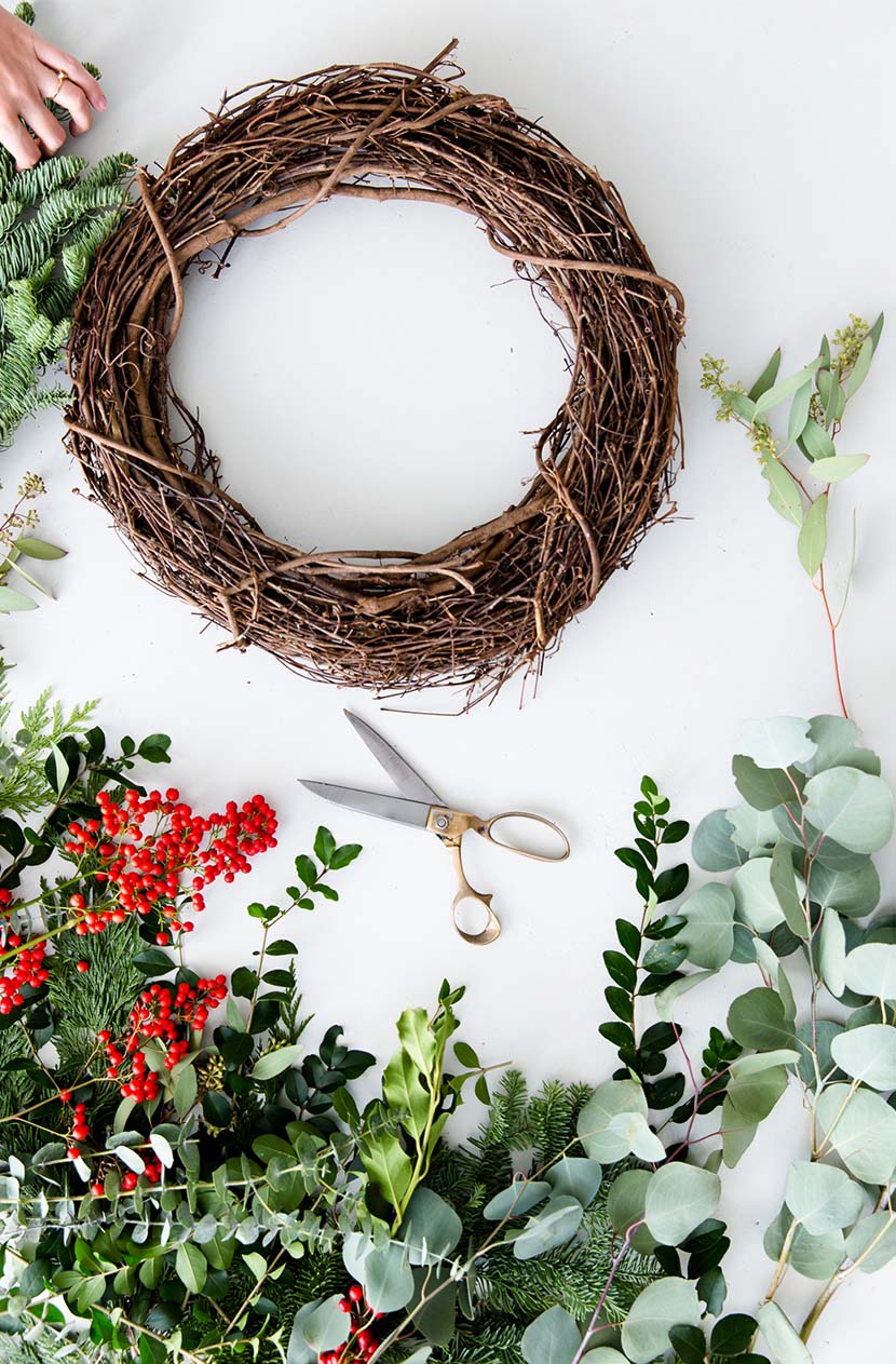 Holiday wreath vines are put together to create a circle clippings of holly branches and tree leaves with scissor on white table.